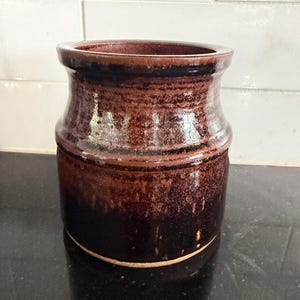 May include: A cylindrical ceramic crock featuring a glossy, deep brown glaze with darker, near-black tones at the base and a subtle horizontal ridge encircling the upper section. The vessel has a wide, open mouth and a smooth, reflective finish.