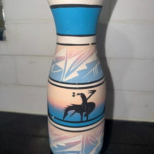 May include: A cylindrical ceramic vase featuring a matte finish with a gradient of soft pink, blue, and cream colors. The vase is decorated with horizontal black bands and etched geometric leaf patterns, along with a black silhouette of a figure.