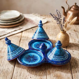 May include: A three-sectioned, blue ceramic serving dish with matching lids. The dish features a floral design in the center of each section and is placed on a wooden surface. A small vase with dried flowers and a stack of plates are in the background.