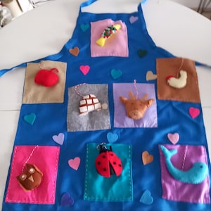 May include: A blue apron with felt appliqués of a red apple, a fish, a turtle, a cow, a ladybug, an owl, a whale, and heart shapes. The apron has blue ties. The appliqués are in various colors.