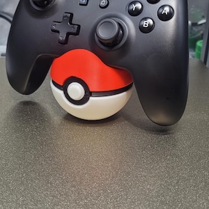 May include: Black Nintendo Switch Pro Controller resting on a red and white Pokeball. The controller has the Nintendo Switch logo and the standard button layout. The Pokeball is a 3D-printed accessory.