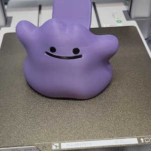 以下が含まれることがあります： ラベンダー色の3DプリントされたDittoの携帯電話スタンド。スタンドは、キャラクターの象徴的なブロブの形と笑顔の顔が特徴です。スタンドは、携帯電話を立てて保持するように設計されています。
