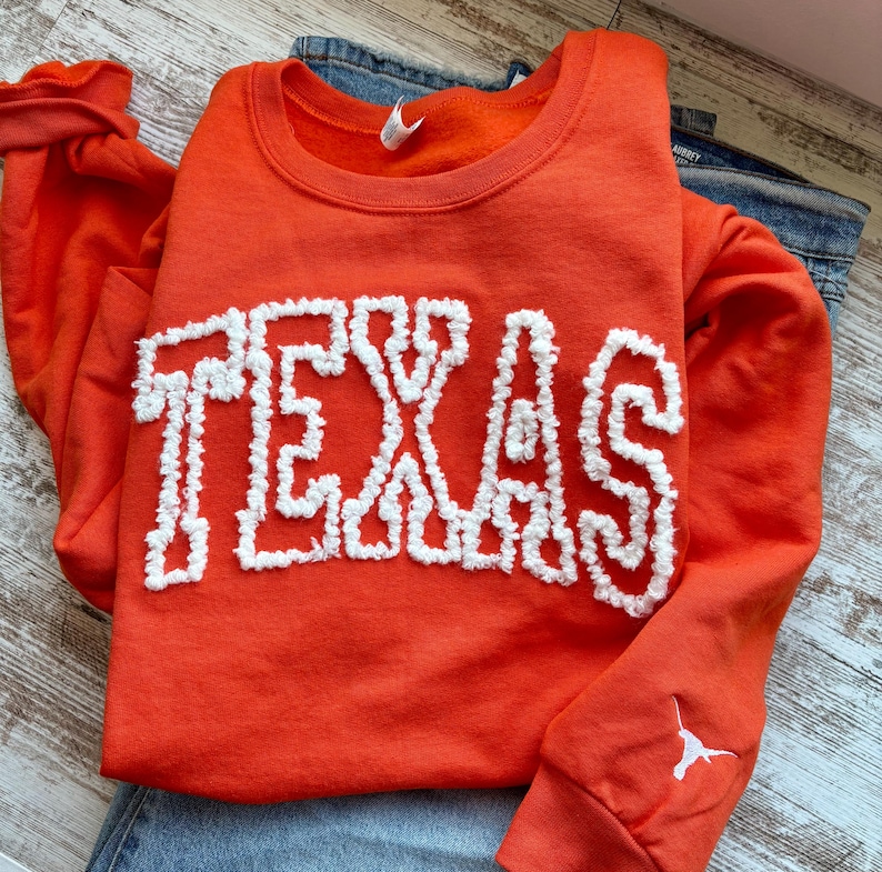 May include: An orange sweatshirt with the word "TEXAS" in white, textured lettering. The sweatshirt has long sleeves and a small white logo on the cuff. The sweatshirt is paired with blue jeans.