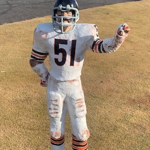 Puede incluir: Una estatua de jugador de fútbol blanco y naranja con casco y camiseta con el número 51. La estatua está de pie sobre una base verde.