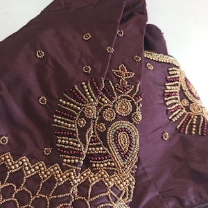 Puede incluir: Una prenda de color burdeos oscuro con intrincados bordados de cuentas doradas y granates. El diseño presenta patrones y adornos detallados, con un enfoque en motivos florales y de cachemira ornamentados. La tela tiene un brillo sutil, realzando la riqueza de los colores.