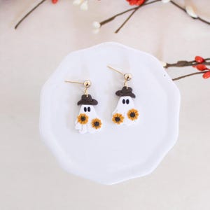 May include: A pair of white ghost earrings with brown hats and yellow sunflower accents. The earrings are on a white plate.