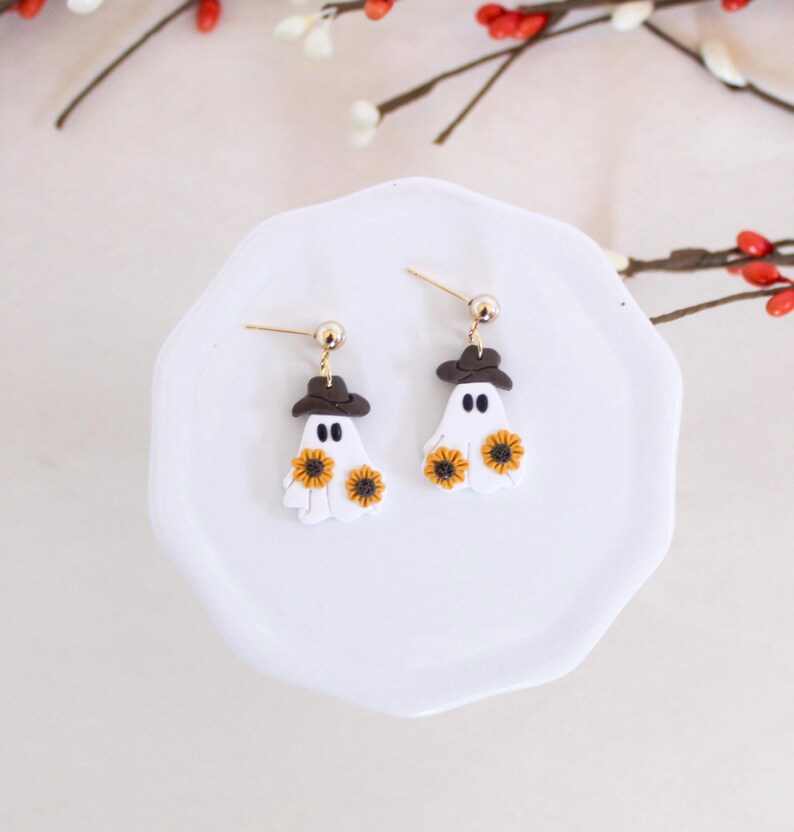 May include: A pair of white ghost earrings with brown hats and yellow sunflower accents. The earrings are on a white plate.