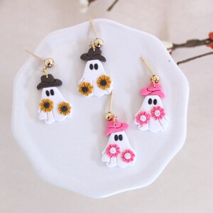 May include: Three pairs of Halloween-themed ghost earrings. Each pair features a white ghost with a different colored hat and flowers. One pair has a brown hat and yellow flowers, another has a pink hat and pink flowers, and the last pair has a pink hat and pink flowers.