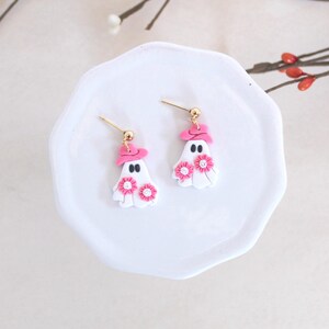 May include: A pair of pink and white ghost earrings with pink flowers and a pink hat. The earrings are on a white plate.