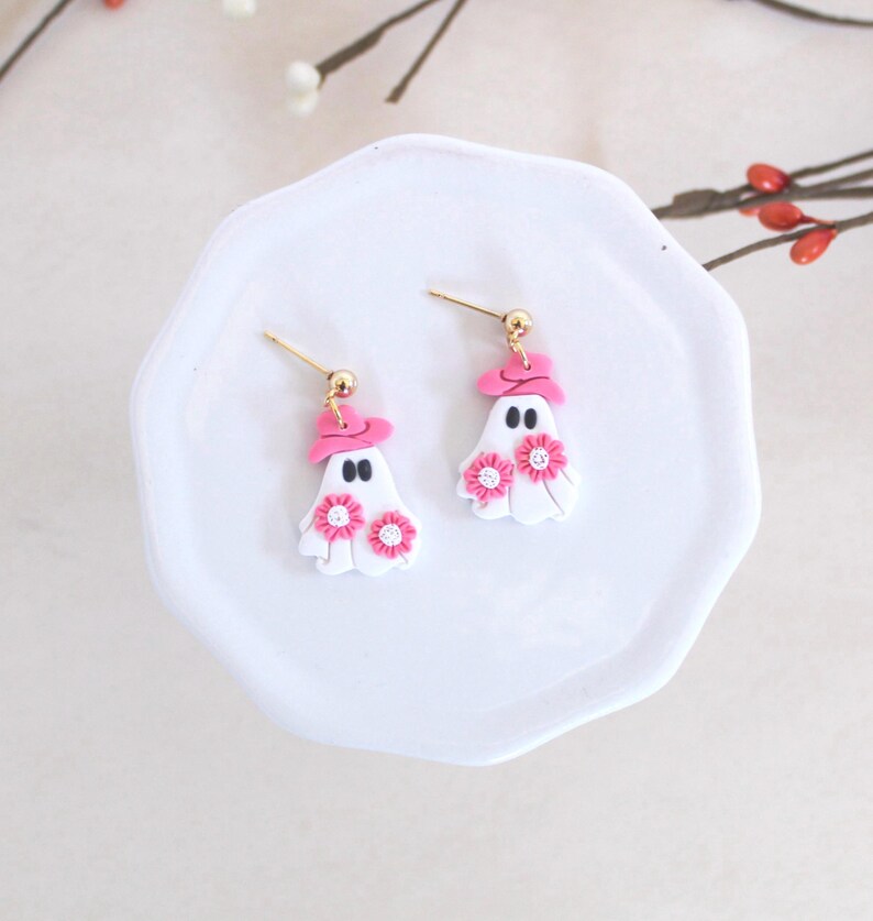 May include: A pair of pink and white ghost earrings with pink flowers and a pink hat. The earrings are on a white plate.