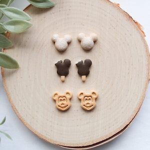 May include: Six pairs of polymer clay earrings on a wooden surface. The earrings are shaped like Mickey Mouse heads and ice cream bars. The Mickey Mouse heads are in white, brown, and tan. The ice cream bars are in brown and white.