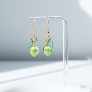 May include: A pair of lemon-shaped earrings. Each earring features a translucent, light green lemon charm with a textured surface and a small green leaf. The charms hang from gold-colored hooks.