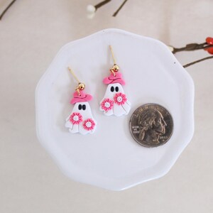 May include: A pair of white ghost earrings with pink hats and pink flowers. The earrings are on a white plate with a US quarter for scale.