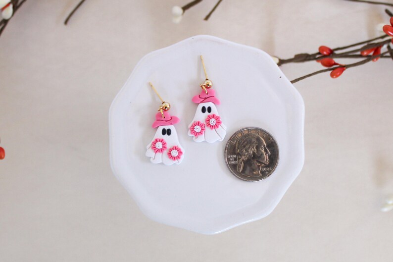 May include: A pair of white ghost earrings with pink hats and pink flowers. The earrings are on a white plate with a US quarter for scale.