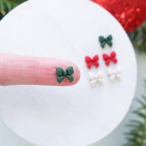 May include: A close-up of a green bow, with other bows in red and white in the background. The bows are small and appear to be made of polymer clay.