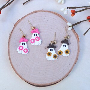 May include: A pair of white ghost earrings with pink and brown hats and flower accents. The earrings are on a wooden surface.