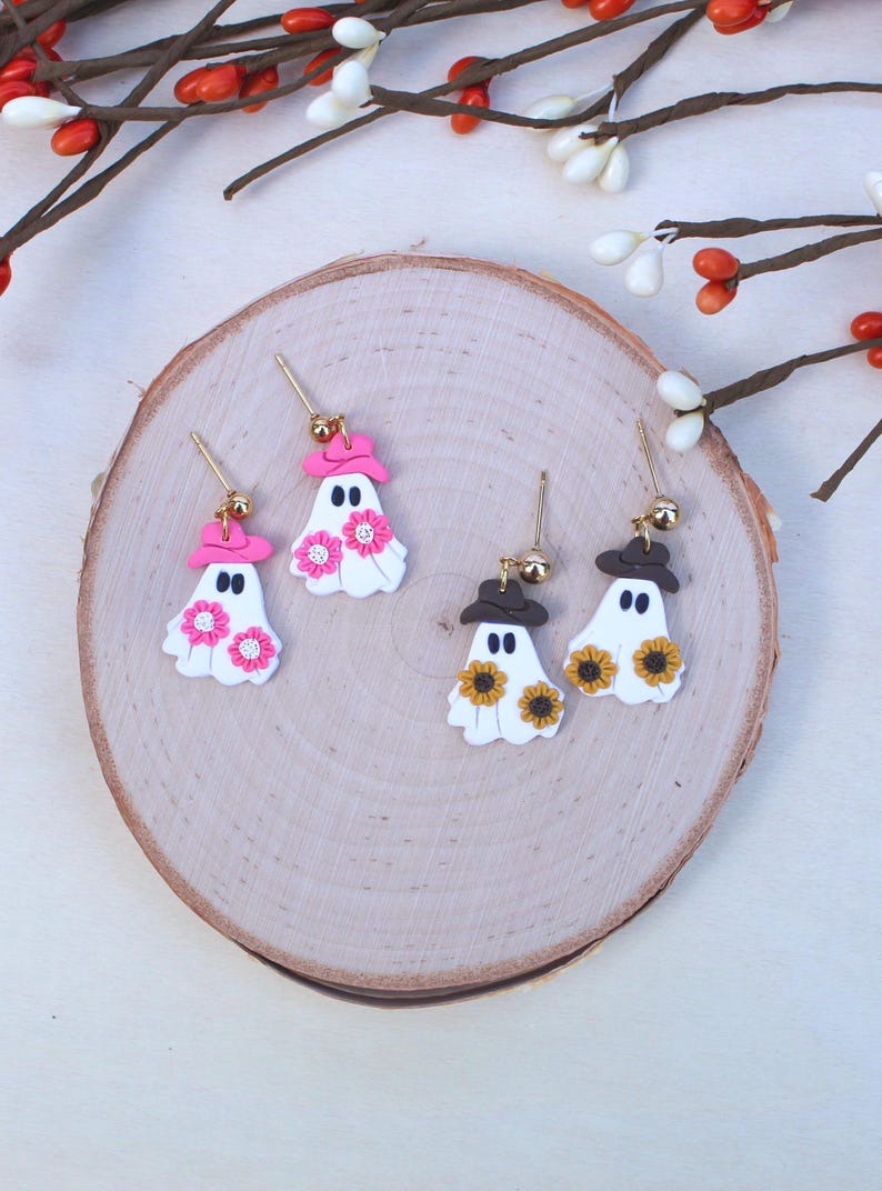 May include: A pair of white ghost earrings with pink and brown hats and flower accents. The earrings are on a wooden surface.