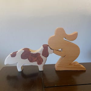 May include: Wooden dog and person figurines. The dog is white with brown spots. The person is kneeling, with the dog appearing to be kissing them. The figurines are made of light-colored wood and are on a dark brown surface.