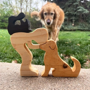 May include: Wooden cutout of a person holding a dog, with a black cutout of a cat on top of the person's head. The cutout is made of light brown wood and is sitting on a gray surface. A golden retriever dog is blurred in the background.