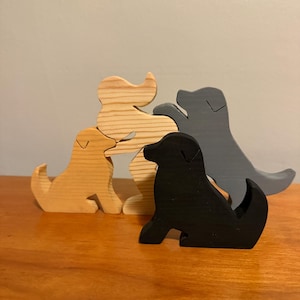 May include: Four wooden dog figurines in various colors: natural wood, tan, black, and gray. The dogs are in a sitting position, with their tails curled. The figurines are arranged on a wooden surface.