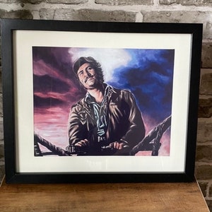 Puede incluir: Impresión enmarcada de un hombre con una chaqueta de cuero marrón mirando hacia arriba a un cielo colorido. El hombre lleva una camisa azul y una gorra. El fondo es un cielo dramático con tonos de rojo, morado y azul.