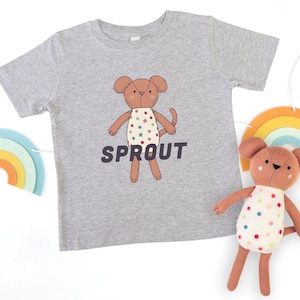 May include: Heather gray t-shirt with a cartoon bear graphic and the word "SPROUT". The bear wears a polka-dot dress. A matching stuffed bear and rainbow decor are also visible.