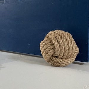 May include: A close-up of a natural fiber rope doorstop in the shape of a knot. The doorstop is beige and is sitting on a white tile floor in front of a blue door.