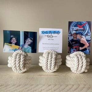 Könnte beinhalten: Drei cremefarbene Seilknoten-Fotohalter auf einer hellen Holzoberfläche. Jeder Halter trägt ein anderes Foto oder eine Karte. Die mittlere Karte trägt die Aufschrift "Knots By Bailz" und "Ask about custom orders!"