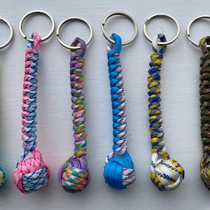 May include: Seven colorful keychains with a knotted ball at the end. The keychains are made of braided paracord in various colors, including blue, green, yellow, pink, purple, and camouflage.