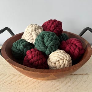May include: A wooden bowl with black metal handles filled with decorative rope balls. The balls are in shades of cream, green, and burgundy. The bowl sits on a light-coloured wooden surface.