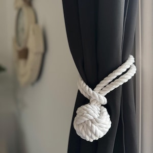 May include: A white nautical rope curtain tieback is knotted around a black curtain. The tieback is made of thick, twisted rope, forming a decorative knot. The background is blurred, showing a wall and a decorative clock.