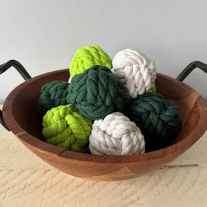 May include: A wooden bowl with black metal handles is filled with decorative rope knots in shades of green and white. The knots are tightly wound and create a textured appearance, perfect for home decor.