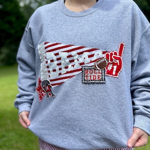 May include: A gray sweatshirt with appliqued patches. The patches include a red and white pennant with "BAMA" in white, a football, a red foam finger with "#1", and a patch that says "ROLL TIDE". A small football player is also on the sweatshirt.