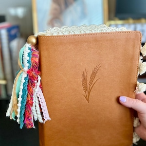May include: A brown leather journal with a wheat stalk design embossed on the cover. The journal has a colorful tassel with gold beads, and white lace trim. A hand is holding the journal.