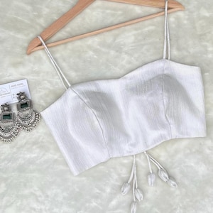 May include: White sleeveless crop top with a white drawstring and tassel detail. The top is made of a textured fabric and has a fitted silhouette.