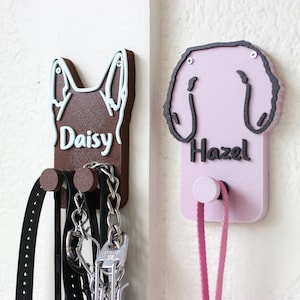 May include: Two wooden dog leash holders, one brown with the name "Daisy" and the other pink with the name "Hazel". Both holders have a hook for hanging leashes.