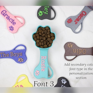 K&ouml;nnte beinhalten: Ein Set aus neun bunten, knochenf&ouml;rmigen Schaufeln f&uuml;r Hunde mit personalisierten Namen. Die Schaufeln sind aus Kunststoff und haben ein Pfotenabdruck-Design. Die Namen auf den Schaufeln sind Gracie, Oscar, ley, Rosie, The Boys, Odin und 12. Die Schaufeln sind in zuf&auml;lliger Reihenfolge auf einer wei&szlig;en Oberfl&auml;che angeordnet. Schriftart 3.