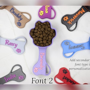 K&ouml;nnte beinhalten: Eine Sammlung von bunten, knochenf&ouml;rmigen Hundefutterl&ouml;ffeln mit Pfotenabdr&uuml;cken und personalisierten Namen, darunter Maverick, Sunny, Brandy, Roxy, Indie, Rocket und Buddy. Die L&ouml;ffel sind aus Kunststoff und haben ein Pfotenabdruckdesign am Griff. Der Text "Font 2" wird unter den L&ouml;ffeln angezeigt.