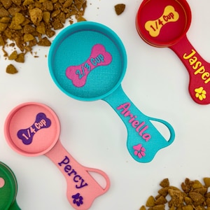 May include: Colorful pet food measuring scoops in teal, pink, red, and green. Each scoop has a bone-shaped measurement indicator and a name. The scoops measure 1/4 cup and 2/3 cup. Dry pet food is scattered around the scoops.