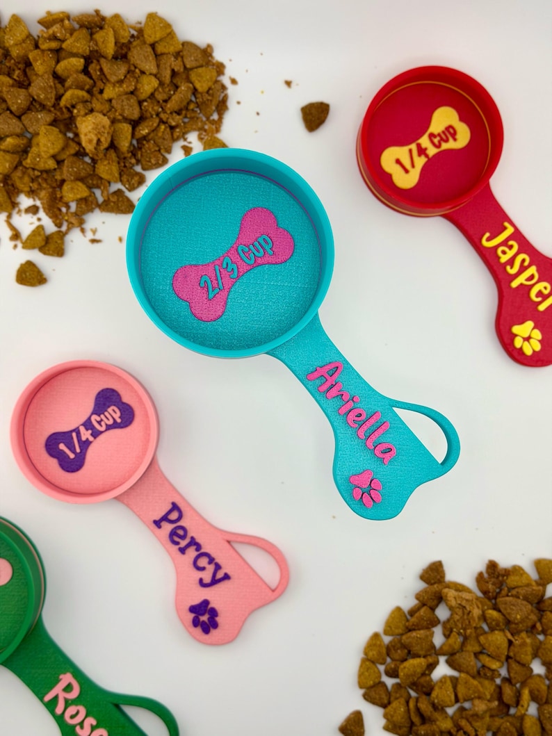 May include: Colorful pet food measuring scoops in teal, pink, red, and green. Each scoop has a bone-shaped measurement indicator and a name. The scoops measure 1/4 cup and 2/3 cup. Dry pet food is scattered around the scoops.