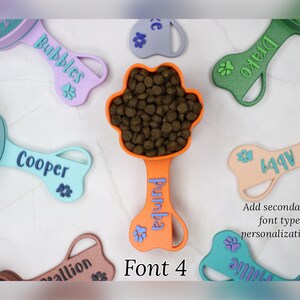 K&ouml;nnte beinhalten: Ein Set aus neun bunten, knochenf&ouml;rmigen Hundefutterl&ouml;ffeln mit personalisierten Namen. Die L&ouml;ffel sind aus Kunststoff und haben ein Pfotenabdruck-Design. Die Namen auf den L&ouml;ffeln sind Bubbles, Drako, Pumba, Cooper, 997, allion und Julie. Der Text "Font 4" wird unter den L&ouml;ffeln angezeigt.