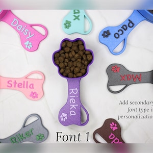 K&ouml;nnte beinhalten: Eine Sammlung bunter, knochenf&ouml;rmiger Hundefutterl&ouml;ffel mit Pfotenabdr&uuml;cken und personalisierten Namen, darunter Daisy, Stella, Rieka, Paco, Riker, Max und da. Die L&ouml;ffel sind aus Kunststoff und haben an einem Ende einen Messbecher. Der Text "Font 1" wird unter den L&ouml;ffeln angezeigt.