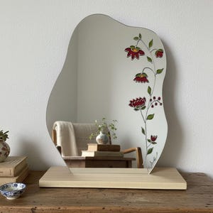 May include: An organically shaped mirror with a floral pattern. The mirror is mounted on a light-coloured wooden base. The floral design includes red flowers and green leaves. The mirror is displayed on a wooden table.