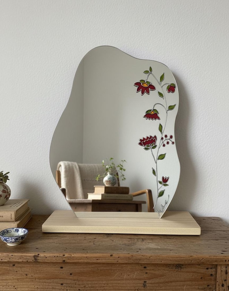 Puede incluir: Espejo de forma irregular con dise&ntilde;o floral. El espejo est&aacute; sostenido por una base de madera de color claro. El dise&ntilde;o floral presenta flores rojas y hojas verdes. El espejo est&aacute; colocado sobre una mesa de madera.