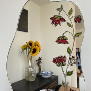 Puede incluir: Un espejo decorativo con una forma ondulada &uacute;nica. El espejo presenta un dise&ntilde;o floral con flores rojas, hojas verdes y un contorno plateado. El espejo refleja un jarr&oacute;n con girasoles y otros objetos.