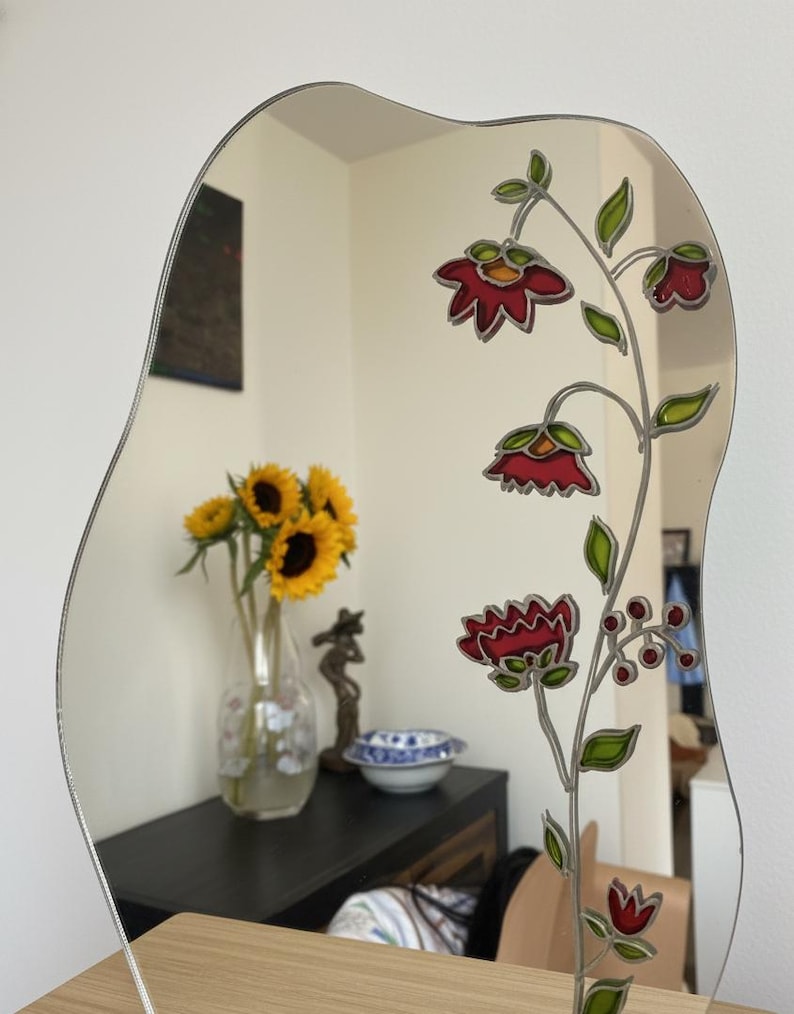 Puede incluir: Un espejo decorativo con una forma ondulada &uacute;nica. El espejo presenta un dise&ntilde;o floral con flores rojas, hojas verdes y un contorno plateado. El espejo refleja un jarr&oacute;n con girasoles y otros objetos.