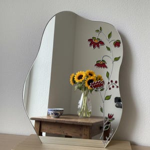 Puede incluir: Espejo decorativo de forma irregular, con un dise&ntilde;o floral en rojo, verde y negro. El espejo est&aacute; montado sobre una base de madera clara. El reflejo muestra un jarr&oacute;n con girasoles y un cuenco peque&ntilde;o.