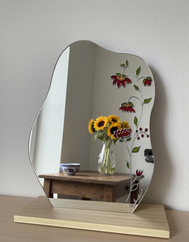 Puede incluir: Espejo decorativo de forma irregular, con un dise&ntilde;o floral en rojo, verde y negro. El espejo est&aacute; montado sobre una base de madera clara. El reflejo muestra un jarr&oacute;n con girasoles y un cuenco peque&ntilde;o.