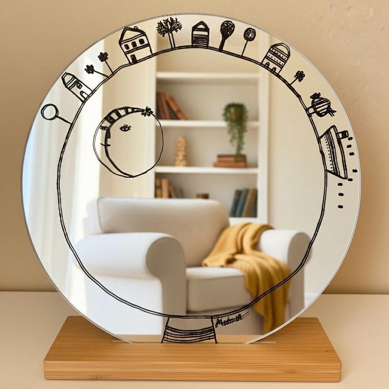 Puede incluir: Un espejo redondo con un dibujo lineal negro de un paisaje caprichoso, que incluye casas, &aacute;rboles y una luna. El espejo est&aacute; montado sobre una base de madera rectangular. El reflejo muestra un sill&oacute;n blanco y una manta amarilla.