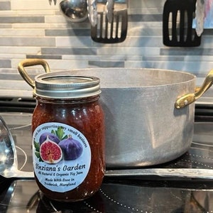 May include: A glass jar of fig jam with a label that reads "Sinziana's Garden All Natural & Organic Fig Jam Made With Love in Frederick, Maryland". The jar is on a black surface next to a silver ladle and a large silver pot with a brass handle.